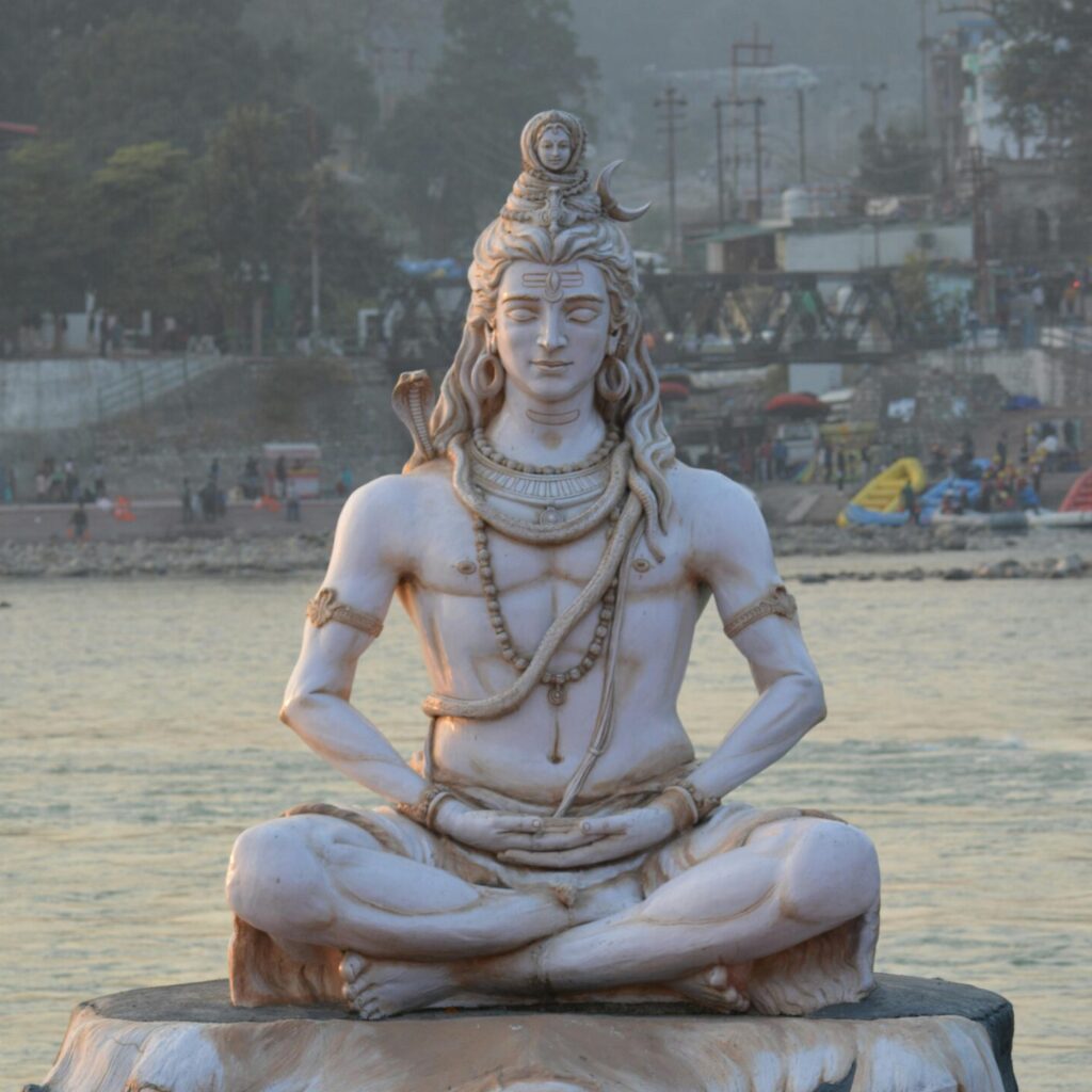 A grand Lord Shiva statue by the Ganges River in Rishikesh, India, surrounded by visitors.
